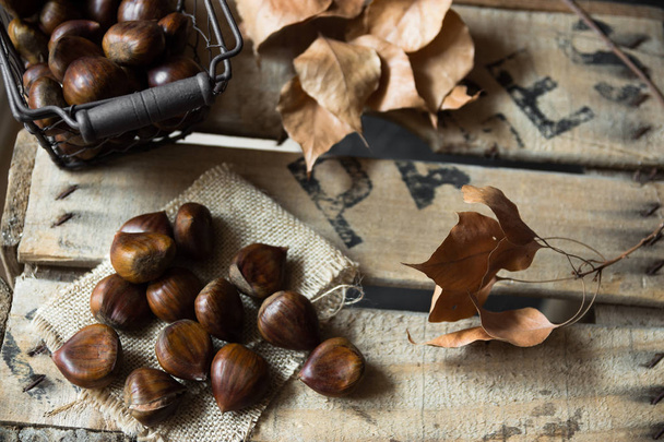 Fresh shiny chestnuts in wire basket scattered on burlap cloth, dry brown leaves,on vintage wood box, autumn, fall, harvest - Photo, Image