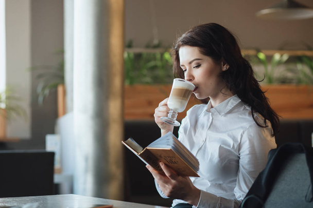 Mujer joven relajándose en la cafetería, bebiendo café con leche, leyendo el libro del diario
 - Foto, Imagen