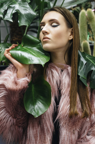 Hermosa mujer joven con grandes hojas verdes en falso abrigo de piel ecológica sobre fondo blanco a cuadros
. - Foto, Imagen