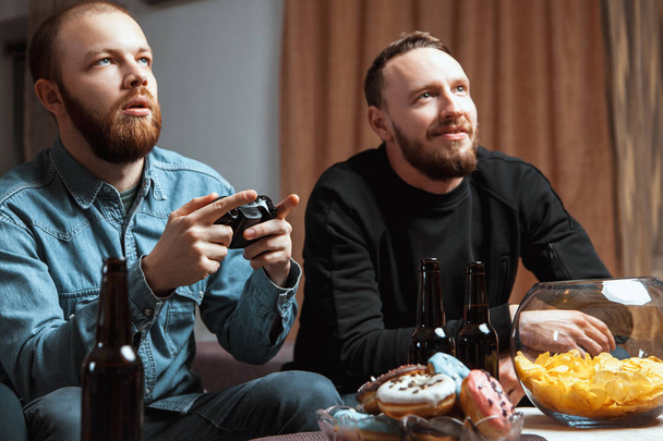 A company of men having fun and laughing sitting on the couch at home with beer and chips, two of them with joysticks in hand playing computer video game - Fotografie, Obrázek