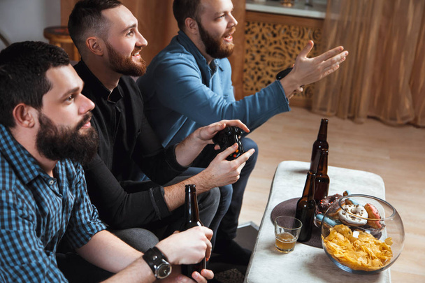 A company of men having fun and laughing sitting on the couch at home with beer and chips, two of them with joysticks in hand playing computer video game - Foto, imagen