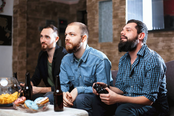 A company of men having fun and laughing sitting on the couch at home with beer and chips, two of them with joysticks in hand playing computer video game - Zdjęcie, obraz