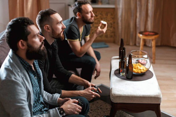 A company of men having fun and laughing sitting on the couch at home with beer and chips, two of them with joysticks in hand playing computer video game - 写真・画像