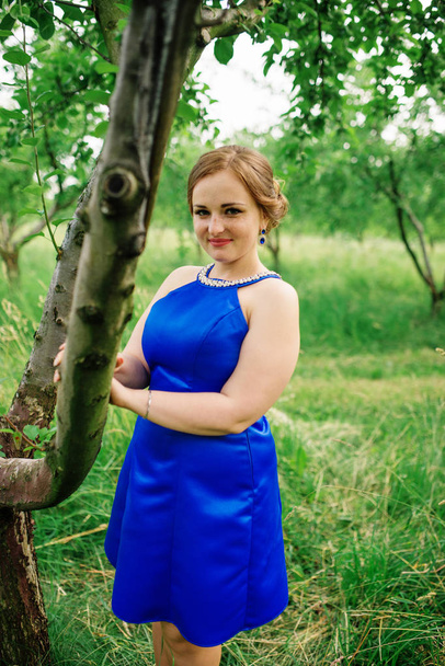 Young overweight girl at blue dress posed background spring gard - Photo, image
