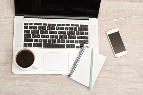 Κούπα του καφέ και ένα σημειωματάριο σε ένα ανοιχτό laptop, smartphone. Ανοιχτόχρωμο φόντο ξύλου, κάτοψη - Φωτογραφία, εικόνα