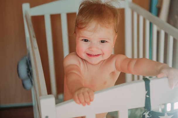 Feliz lindo bebé sonriendo con los dientes y sentado en la cuna, acaba de despertar
 - Foto, Imagen