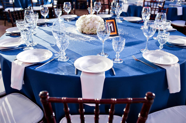 Mariage. Un banquet. Les chaises et la table ronde pour les invités, servis avec couverts et vaisselle et recouverts d'une nappe bleue
. - Photo, image