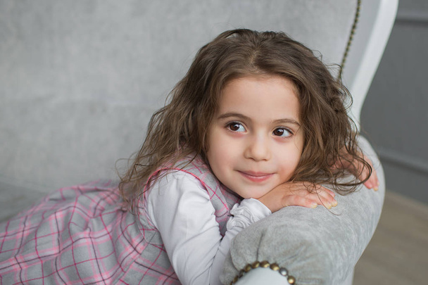 Pretty little girl with brown hair sits on a grey sofa and looks away - Foto, Imagen