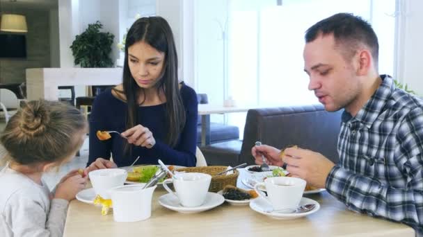 Young lovely parents feeding their blond daughter vegetable salad in cafe. - Felvétel, videó