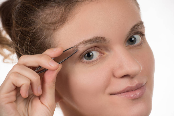 Young girl pulls out her eyebrows with tweezers metal on a white - 写真・画像