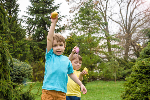 Τα παιδιά στο Πασχαλινό αυγό hunt στην ανθοφορία την άνοιξη στον κήπο. Τα παιδιά ψάχνουν για πολύχρωμα αυγά σε λουλούδι Λιβάδι. Μικρό παιδί αγόρι και ο φίλος του ο αδελφός παιδί παίζουν σε εξωτερικούς χώρους - Φωτογραφία, εικόνα