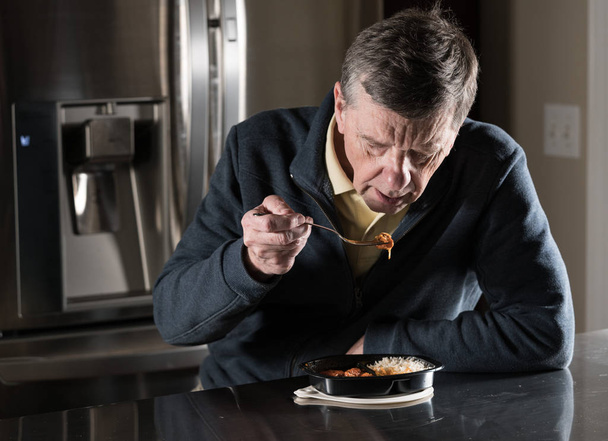 Yksinäinen vanhempi mies syö valmisaterian pöydässä
 - Valokuva, kuva