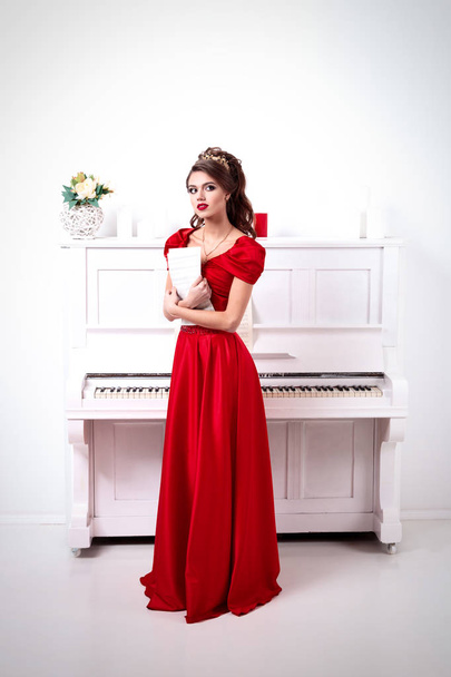 Elegant woman in a long red dress holds unrecognized notes, standing at the piano in a white room - Foto, afbeelding