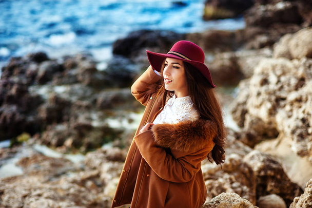 Cheerful lovely young woman in maroon felt hat and drape coat standing and laughing on pier in spring - Foto, imagen
