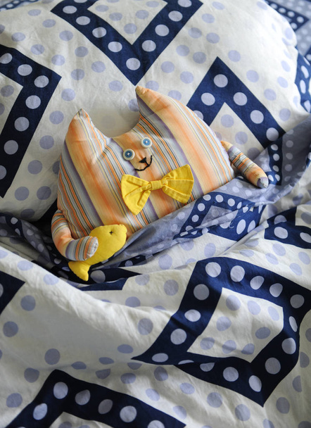 Μαλακό παιχνίδι μια χαριτωμένη γάτα σε ένα κίτρινο παπιγιόν σε μια αγκαλιά με τα ψάρια που είναι ξαπλωμένος σε ένα κρεβάτι που καλύπτεται με μια κουβέρτα - Φωτογραφία, εικόνα