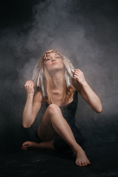 Mujer perfecta vestida de indios americanos en humo sobre un fondo gris. Un sombrero hecho de plumas. Misteriosa manera mística, cuerpo sexy, hermosa espalda. Rubia atractiva con una cara hermosa
 - Foto, Imagen