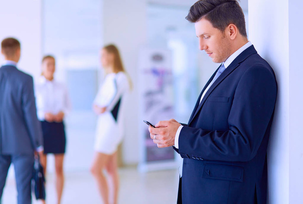 Portrait of young businessman in office with colleagues in the background and using mobile - Photo, image