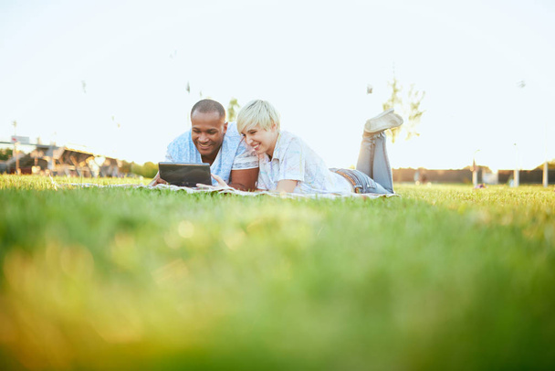 Mixed race couple of millennial in a grass field looking at a digital tablet and reading for their next school paper - Photo, Image