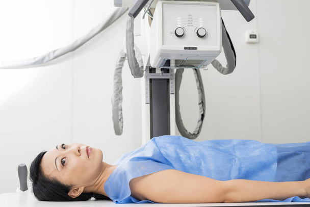 Θηλυκός ασθενής ξαπλωμένος κάτω από X-ray συσκευή στην αίθουσα εξετάσεων - Φωτογραφία, εικόνα