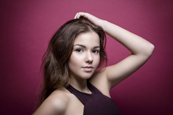 Beautiful and young girl looking at the camera, brunette. The purple top. Gentle make-up, brown eyes, brown hair. On a pink background. Put his hand to his head, he stands sideways. - Foto, imagen