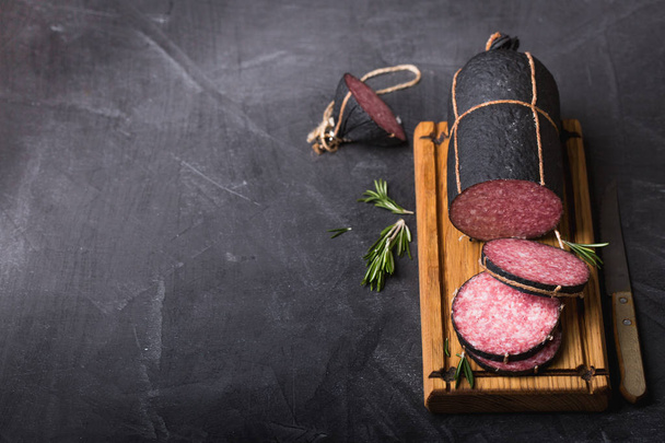 Closeup sausage, sliced on the black coated Board with herbs, thyme, rosemary. Vintage knife, vintage cutting Board. - 写真・画像