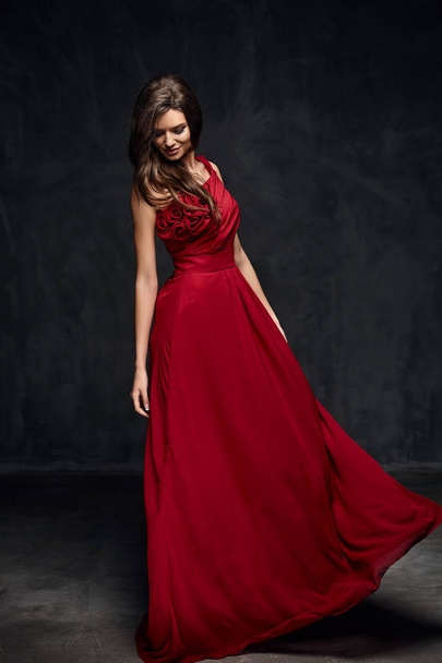 Beautiful young model female with dark hair in long red dress standing in dark studio - Photo, Image