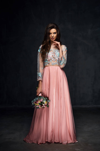 Beautiful young sexy model female with dark hair in gorgeous romantic glossy long light pink dress posing with flowers in dark studio - Zdjęcie, obraz