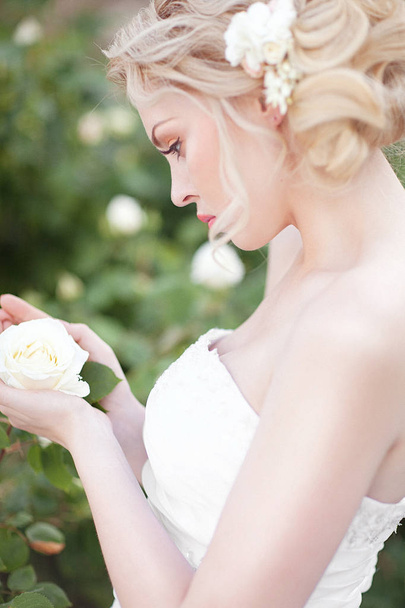Beautiful Brunette Bride near a Flowering Bush Roses Posing in a Wedding Dress - Fotó, kép