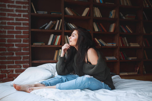 Sensual woman with dark hair in green sweater and jeans sitting on a bed at home - Фото, зображення