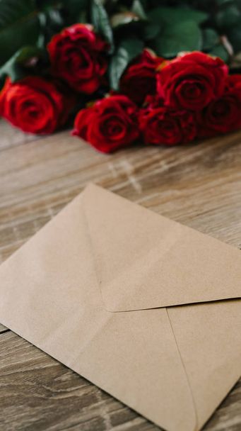 Bouquet of red roses on a wooden table with an envelope and a blank paper card for your text - Foto, afbeelding