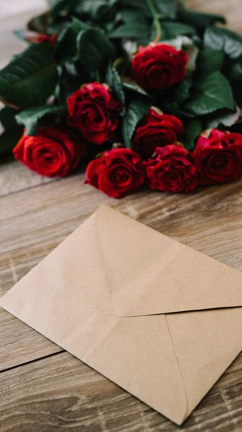 Bouquet of red roses on a wooden table with an envelope and a blank paper card for your text - Foto, afbeelding