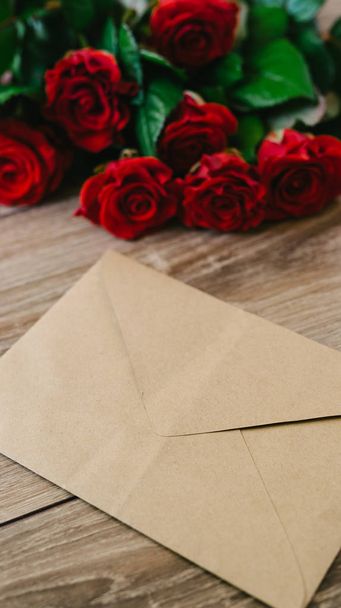 Bouquet of red roses on a wooden table with an envelope and a blank paper card for your text - Fotó, kép