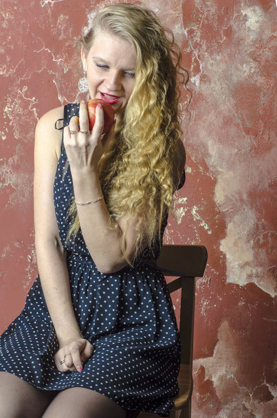 Young blonde girl with curly long hair in a polka-dot dress eating an apple - Fotó, kép