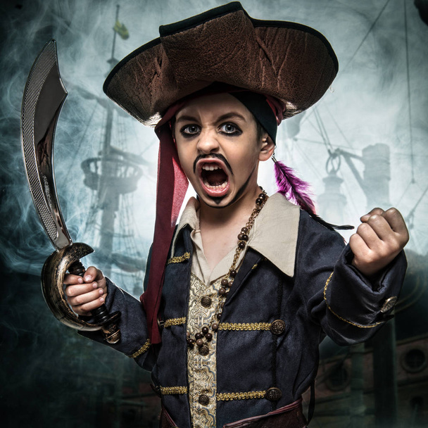 Θυμωμένος νεαρό αγόρι φορώντας ένα κοστούμι πειρατών. Στέκεται το ιστορικό του πλοίου  - Φωτογραφία, εικόνα