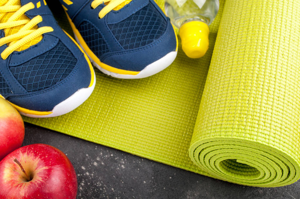 Mata do jogi, buty sportowe, jabłka, butelka wody na ciemnym tle. Pojęcie zdrowego stylu życia, zdrowego odżywiania, sport i dieta. Sprzęt sportowy - Zdjęcie, obraz