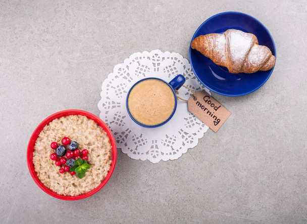 Koffiekopje, havermout met bessen, croissant en Opmerking Goedemorgen op grijze achtergrond. Gezond ontbijt concept - Foto, afbeelding