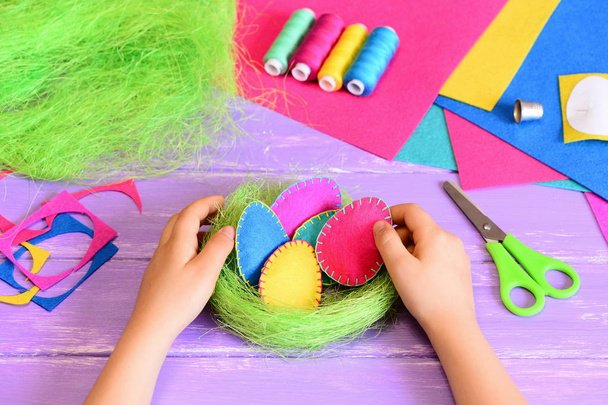 Little child makes Easter decoration. Child puts felt Easter egg in a basket. Beautiful felt Easter eggs and sisal nest decoration, handicraft tools and materials on a table. Easy and nice Easter gift - Photo, Image