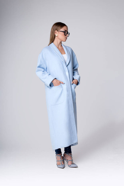 Fashionable blonde in a light blue coat. Photography in full height in a studio on a gray background - Foto, afbeelding
