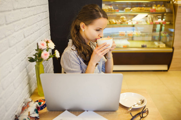 Young girl sitting at table and looking into laptop monitor with cup of drink - Фото, зображення