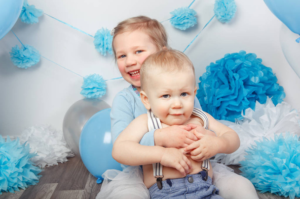 Portrait of two smiling laughing hugging cute adorable Caucasian children, toddler girl and baby boy, celebrating birthday  looking in camera, white studio background with blue balloons - Photo, Image