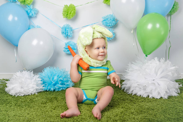 Retrato de lindo adorable bebé caucásico en vacaciones traje de conejo de Pascua con orejas grandes, vestido con ropa verde onesie, sentado en la alfombra suave alfombra esponjosa en el estudio, sosteniendo zanahoria roja
 - Foto, imagen