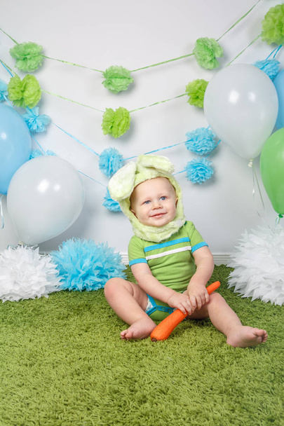 Portrait of cute adorable Caucasian baby boy in holiday Easter bunny rabbit costume with large ears,  dressed in green clothes onesie, sitting on soft fluffy rug carpet in studio, holding red carrot - Photo, Image