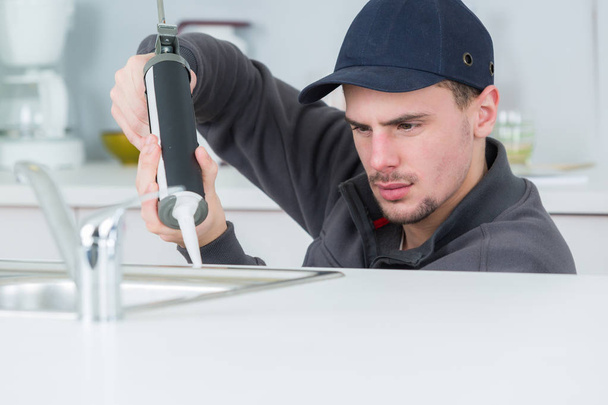 young handyman applying silicone sealant with gun around sink - Foto, afbeelding