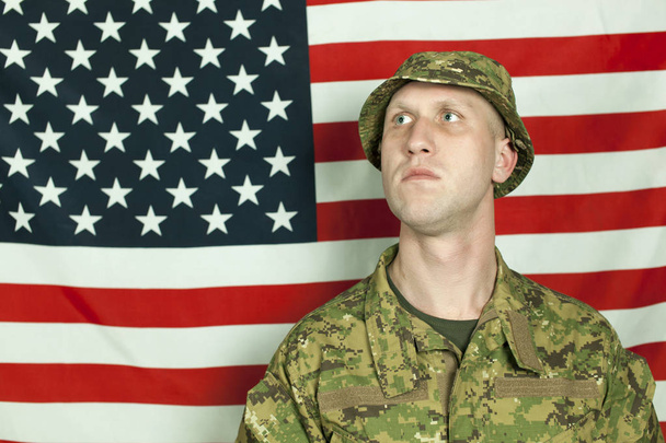 Serios jeune soldat en camouflage debout contre le drapeau américain, regardant de côté.
  - Photo, image