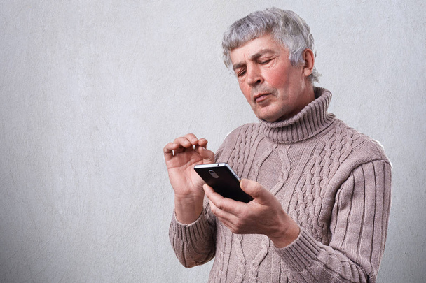 A serious mature man holding smartphone in his hand looking attentively into the screen.  Senior man reading text message on phone having frowned expression. People, technology, lifestyle. - Photo, Image