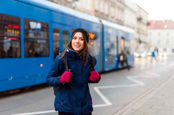 Девушка с рюкзаком улыбается на трамвайной станции
 - Фото, изображение