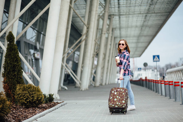 Concepto de viaje aéreo con joven mujer de negocios hipster en camisa a cuadros con maleta y taza desechable con café o té. Libertad de viaje y concepto de vacaciones
. - Foto, Imagen