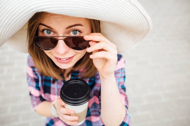 Κάτοψη του μοντέρνα τουριστικά γυναίκα με καπέλο με γυαλιά ηλίου κρατώντας διαθέσιμου Κύπελλο και βλέπουν τα φωτογραφικών μηχανών. - Φωτογραφία, εικόνα