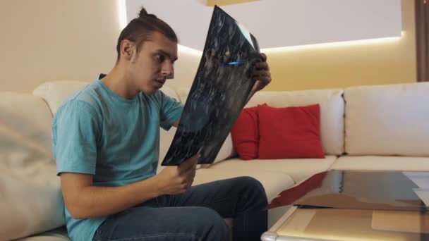 Worried guy in blue shirt sitting on leather sofa look at printed x-ray sheet - Filmagem, Vídeo