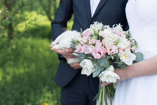 Νύφη που κρατάει στα χέρια της μια λεπτή γαμήλια ανθοδέσμη με λευκά και ροζ τουλίπες και ροζ μικρά τριαντάφυλλα. Γαμπρού κρατώντας ένα λευκό χαριτωμένο κουνέλι.  - Φωτογραφία, εικόνα